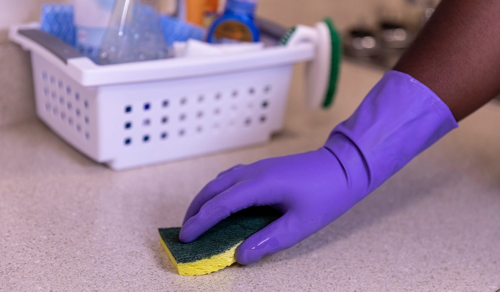 Gloved hand wiping down a kitchen counter with a sponge and cleaning supplies nearby